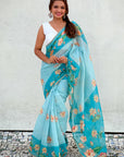 Woman wearing a light blue saree with floral patterns against a white wall.