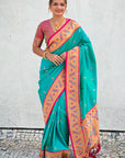 Woman wearing a teal saree with pink and orange border against a light gray wall.