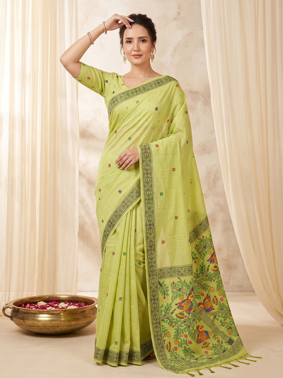 Woman wearing a light green saree with a floral border against a beige curtain background