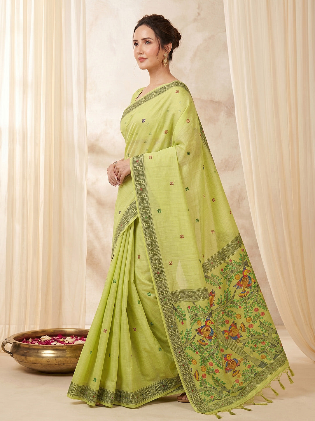 Woman wearing a light green saree with a floral border against a beige curtain background