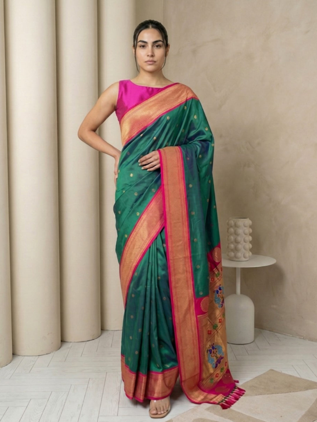 Woman wearing a green and pink saree in a room with beige curtains and a white table.