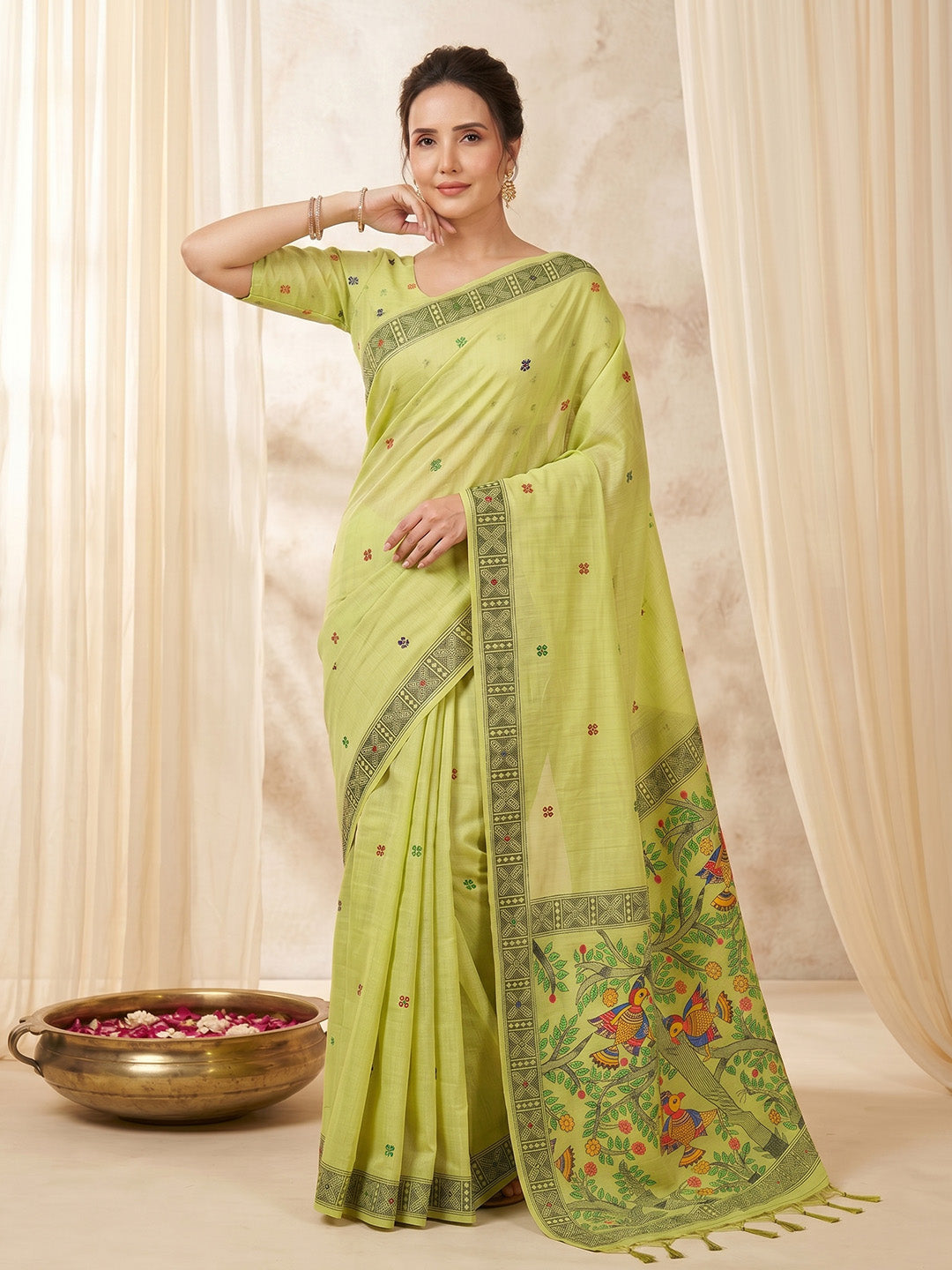 Woman wearing a light green saree with a floral border against a beige curtain background
