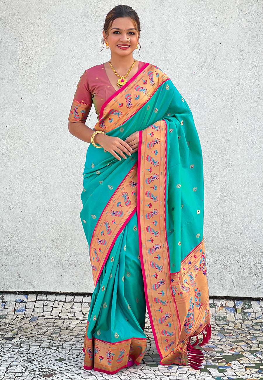 Woman wearing a teal saree with pink and orange border against a light gray wall.